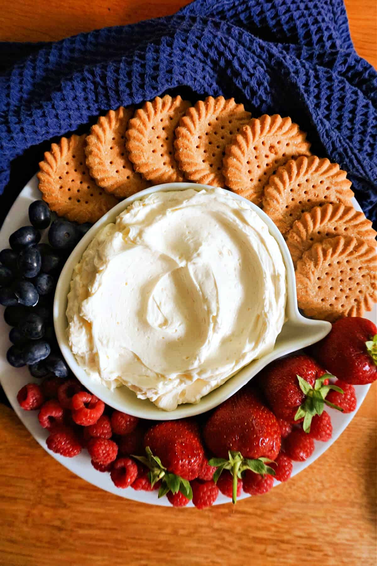 A white plate with a small plate with white dip, round biscuits, and berries around, and a navy blue tea towel around the plate.