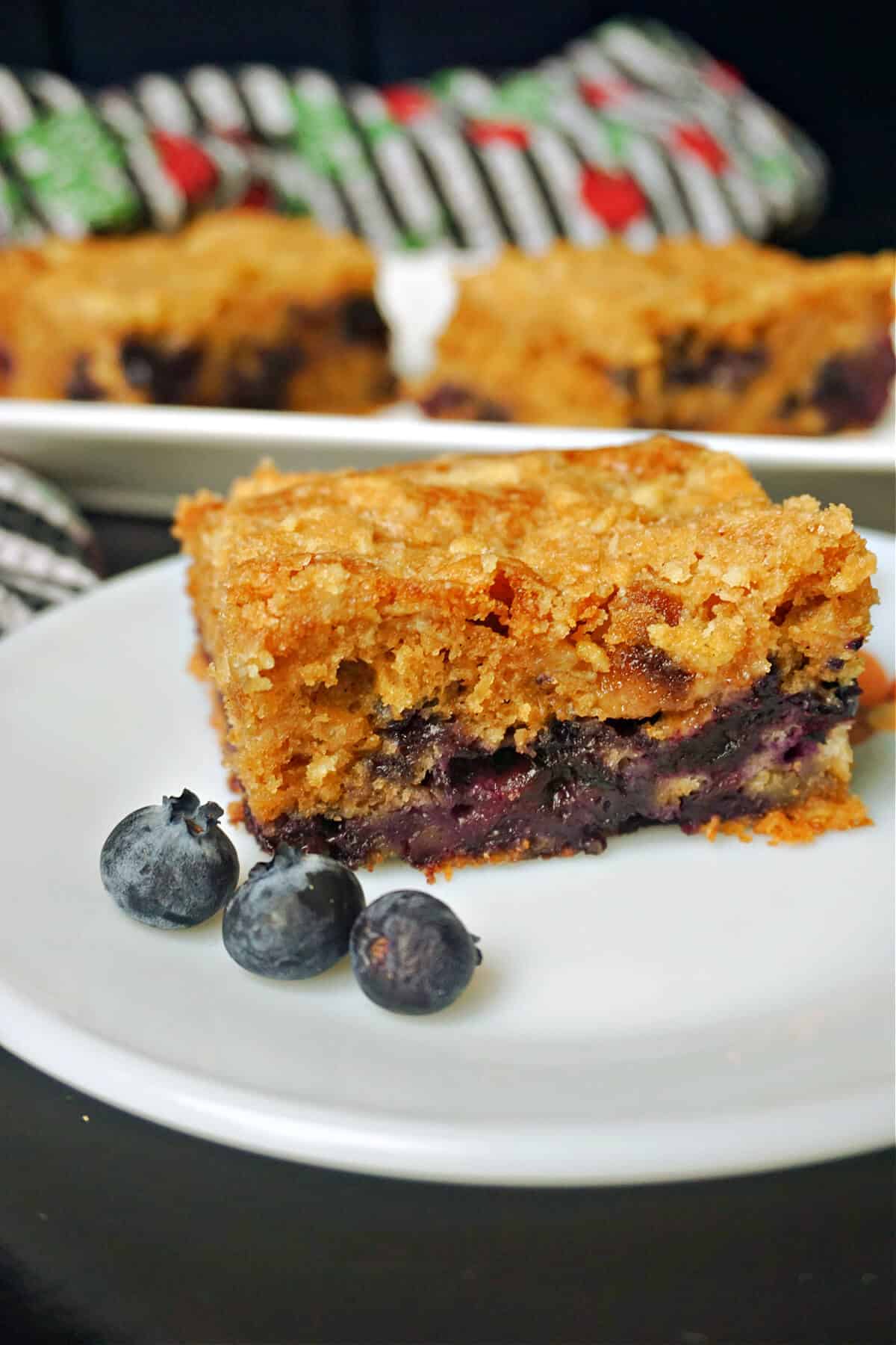 A slice of banana and blueberry cake and 3 blueberries on a white plate with a plate with 2 more slices in the background.
