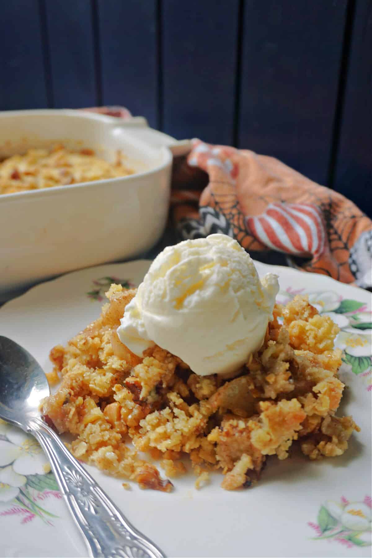 A plate with a teaspoon and a portion of pear crumble and a scoop of ice cream on top. A dish with crumble in the background.