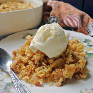 A plate with a teaspoon and a portion of pear crumble topped with a scoop of ice cream. In the background a patterned tea towel and a partiall visible dish with more crumble.