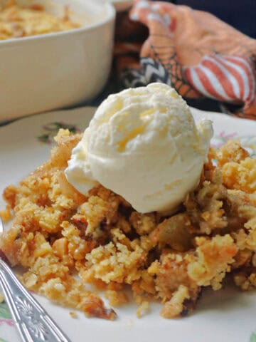 A plate with a teaspoon and a portion of pear crumble topped with a scoop of ice cream. In the background a patterned tea towel and a partiall visible dish with more crumble.