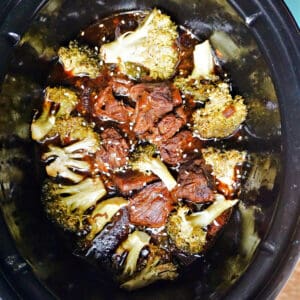 A slow cooker with beef and broccoli florets cooked in sauce and garnished with sesame seeds.