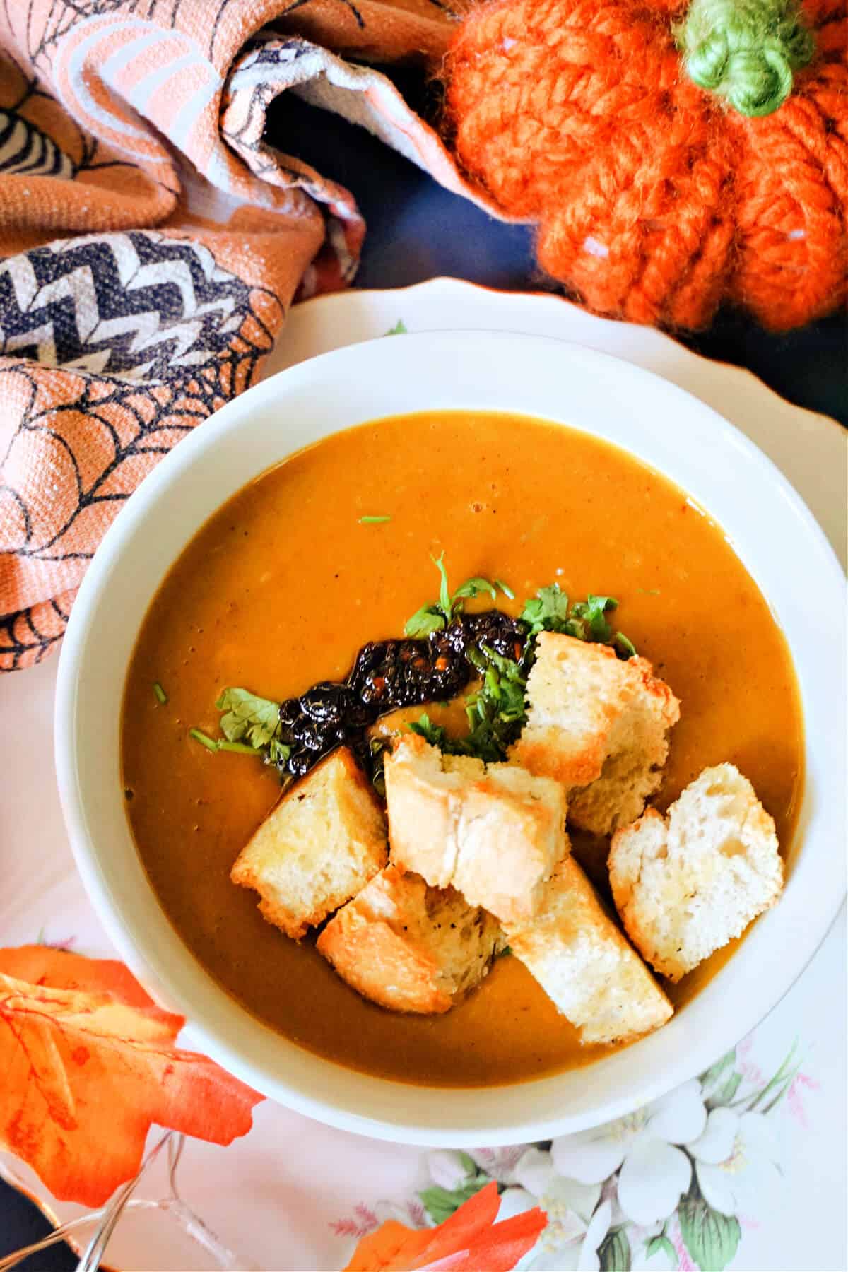 A white bowl with butternut squash soup topped with chopped parsley, chili oil and croutons. Autumnal decorations around it.