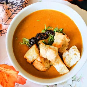 A white bowl with butternut squash soup topped with chopped parsley, chili oil and croutons. Autumnal decorations around it.