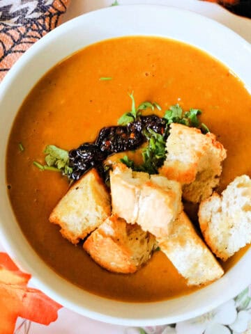 A white bowl with butternut squash soup topped with chopped parsley, chili oil and croutons. Autumnal decorations around it.