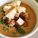 A white bowl with creamy soup topped with croutons, chopped parsley, parmesan and cubed cooked meat.