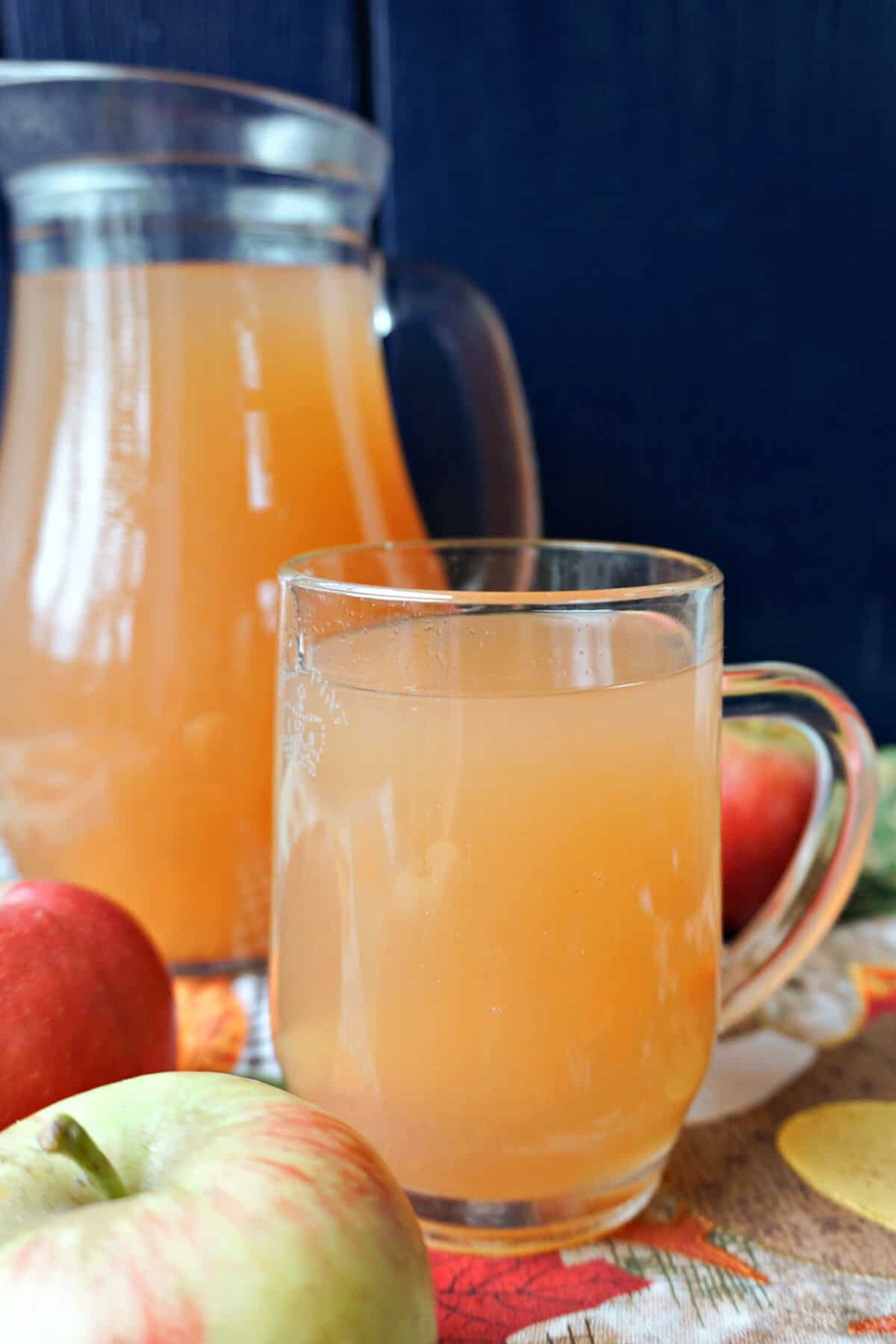 A glass with cider with a jug partially visible in the background and apples around.