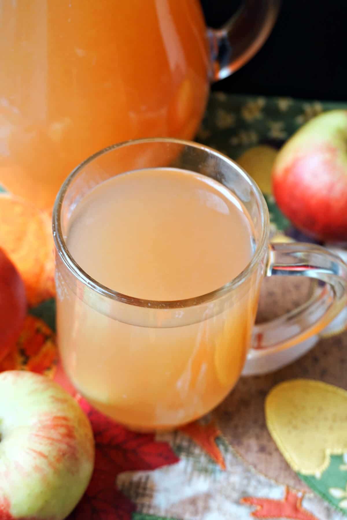 A glass of apple cider with apples around it. A partially-visible jug of cider next to it.