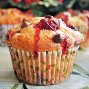 A cranberry choc chip muffin in a muffin case, 2 partially-visible muffins in the background, and Christmas decorations around.