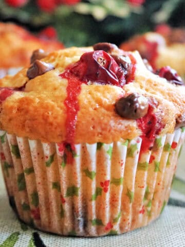A cranberry choc chip muffin in a muffin case, 2 partially-visible muffins in the background, and Christmas decorations around.