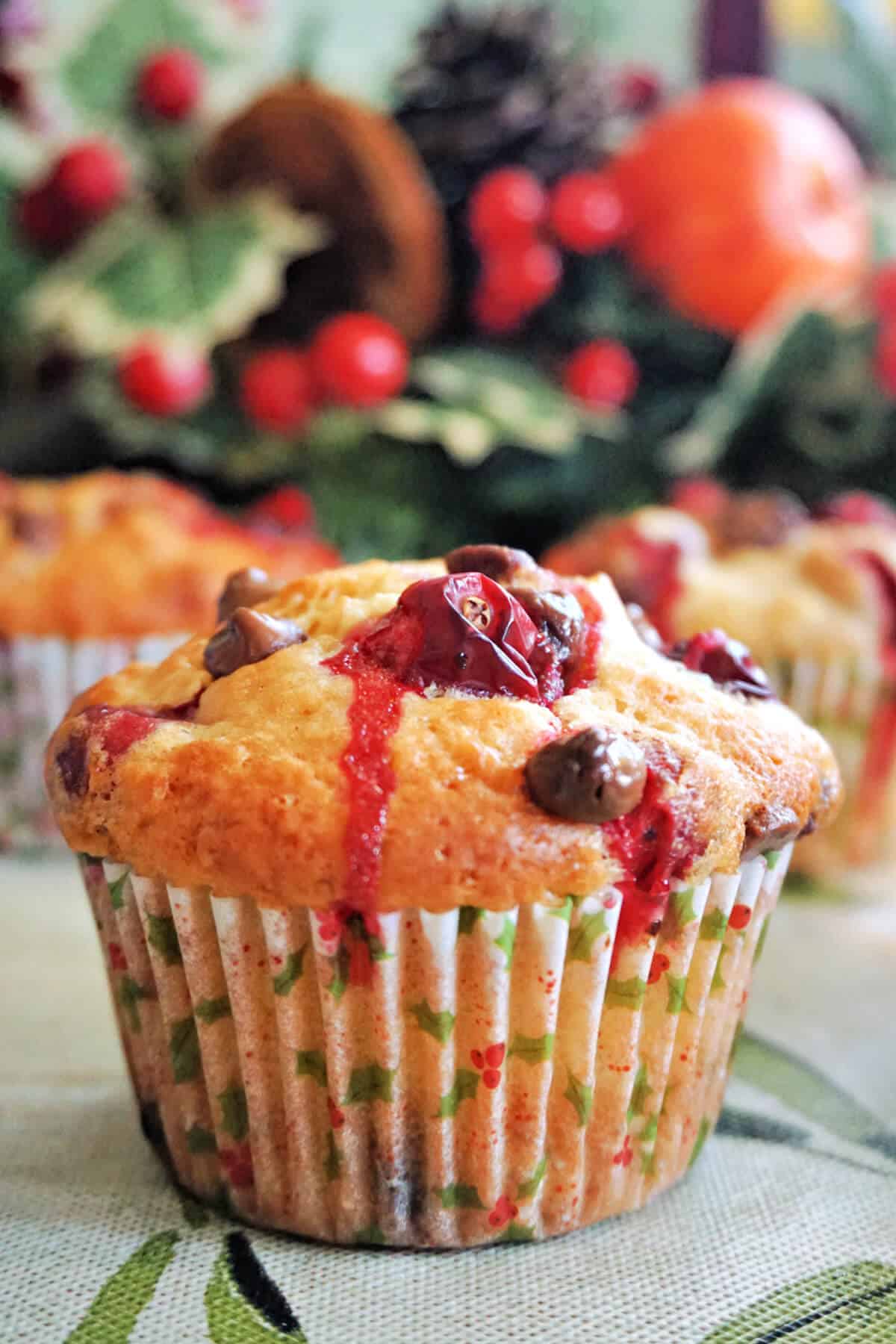 A muffin with cranberries and chocolate chips, more partially visible muffins and Christmas decorations in the background.