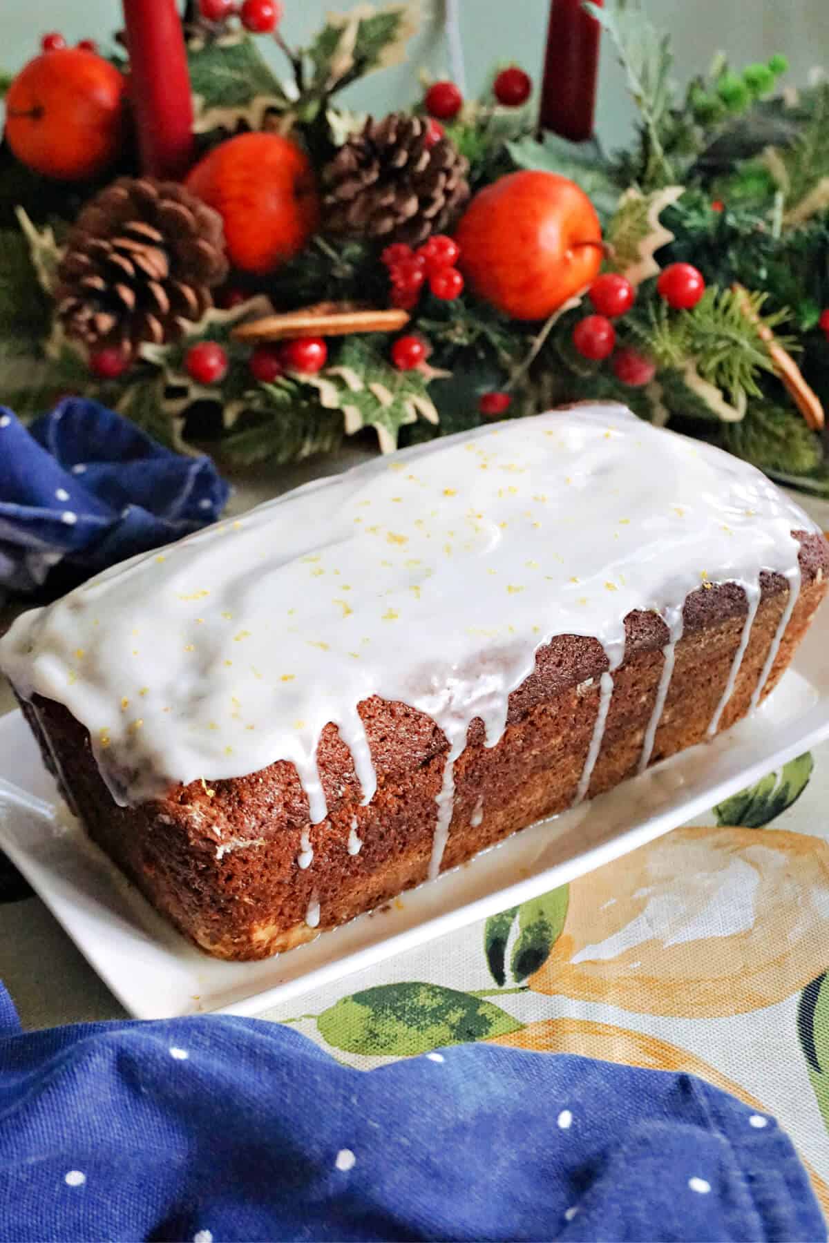 A gingerbread loaf toped with icing on a white rectangle plate with Christmas decorations around.