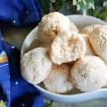 A white bowl with a half eaten almond cookie sitting on top of other whole cookies. Christmas decorations around.