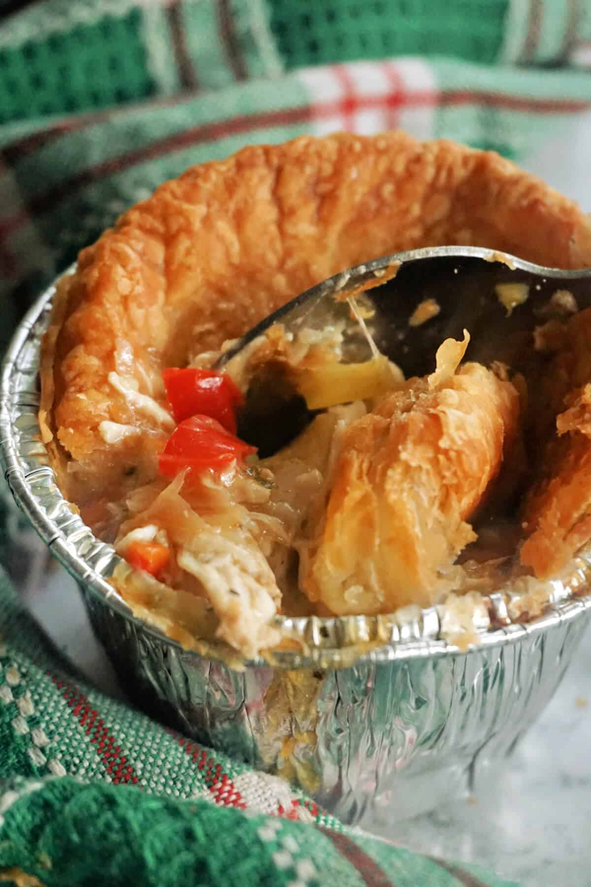 A foil ramekin with a mini chicken pie and a spoon digging into it.