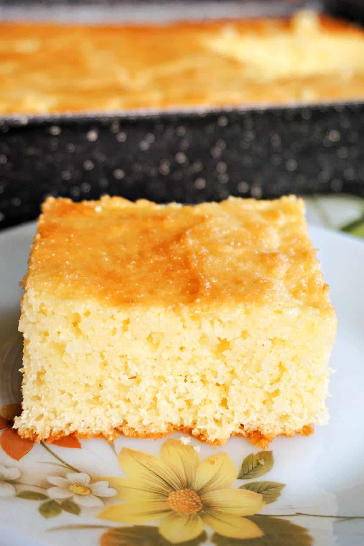 A slice of sponge cake on a plate with a tray of cake in the background.
