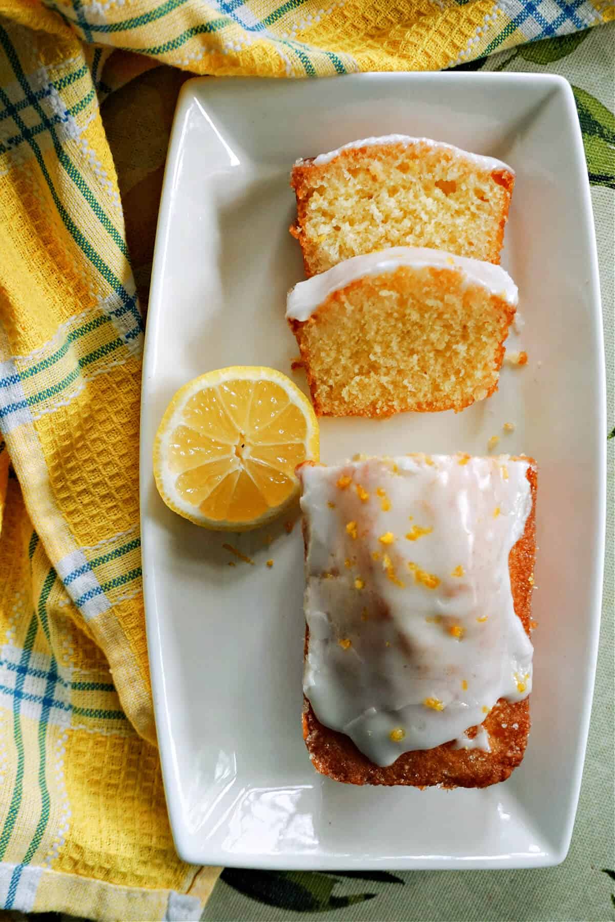 A white rectangle plate with half a lemon, 2 slices of lemon cake and the rst of the iced cake garnished with lemon zest. A yellow-pattered tea towel around the plate.