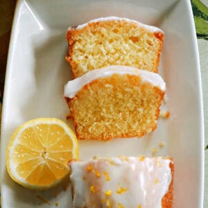 A white rectangle plate with half a lemon, 2 slices of lemon cake and the rst of the iced cake garnished with lemon zest. A yellow-pattered tea towel around the plate.