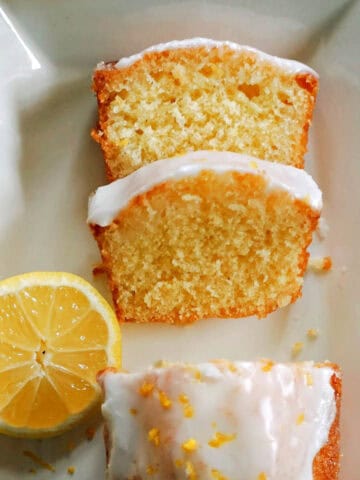 A white rectangle plate with half a lemon, 2 slices of lemon cake and the rst of the iced cake garnished with lemon zest. A yellow-pattered tea towel around the plate.