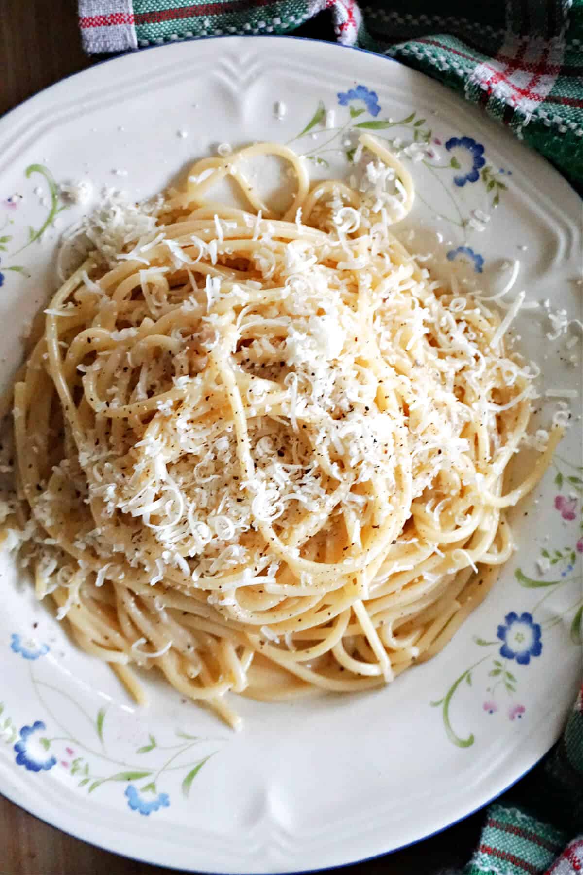 A white flowery plate with a portion of spaghetti garnished with grated cheese and ground black pepper.