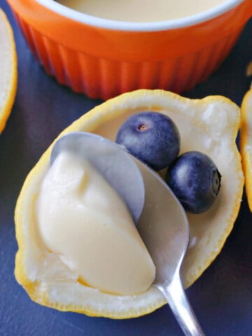 A lemon shell filled with a set cream being scooped out with a teaspoon, topped with 2 blueberries. Other lemon shells and a ramekin filled with cream and topped with blueberries are partially visible.