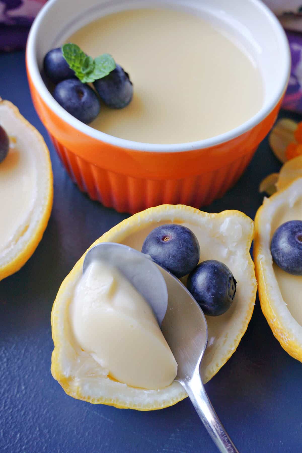 The focus is half a lemon filled with lemon posset being scooped out with a teaspoon. 2 blueberries are resting on top of the posset. Other partially-visible lemon shells and a ramekin filled with posset and topped with 2 blueberries each.