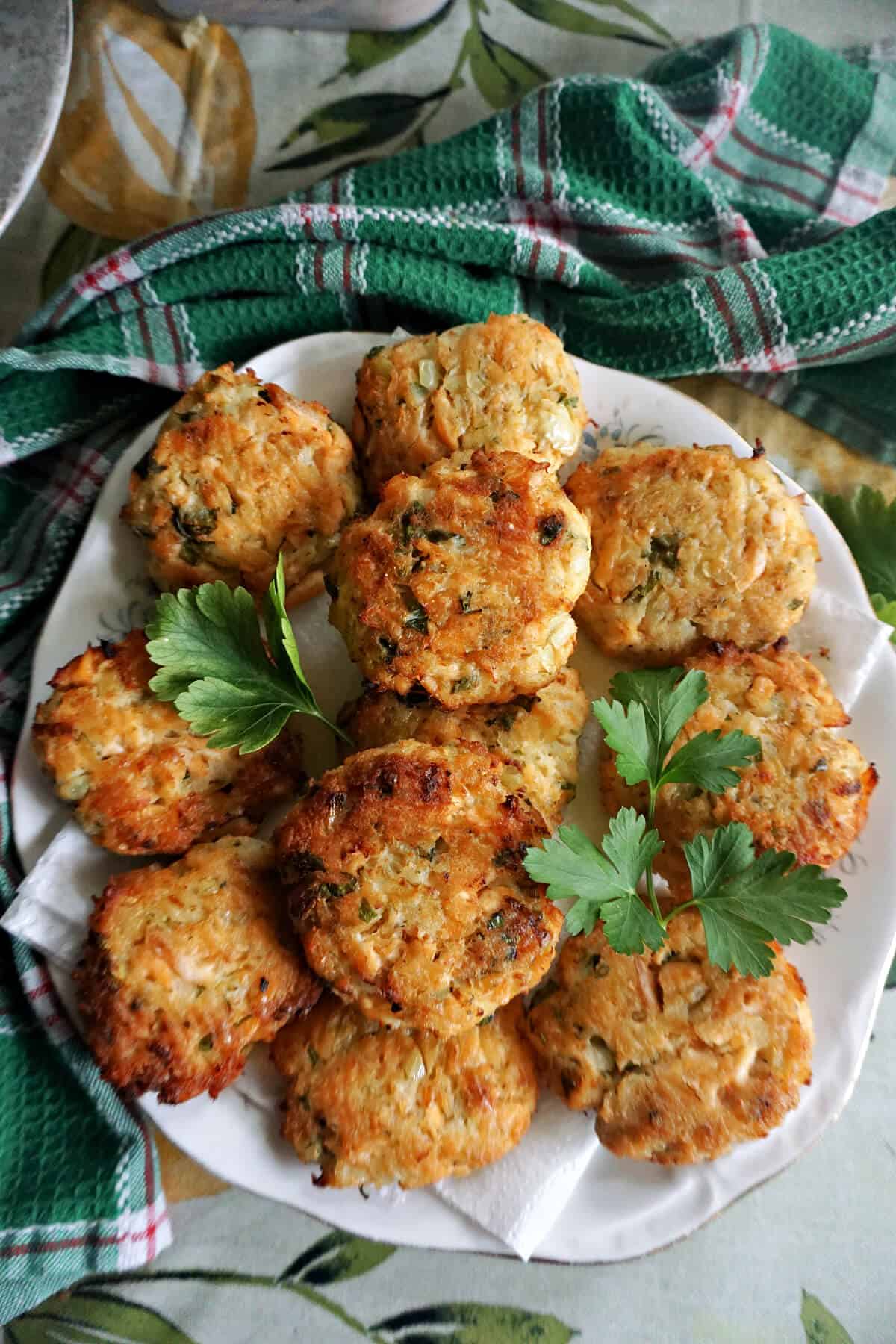 A white plate with salmon patties garnished with fresh parsley leaves and a green tea towel around.