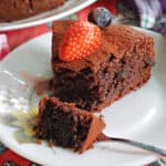 A white flowery plate with a slice of chocolate cake topped with half a strawberry and a blueberry. A fork cutting through the slice.