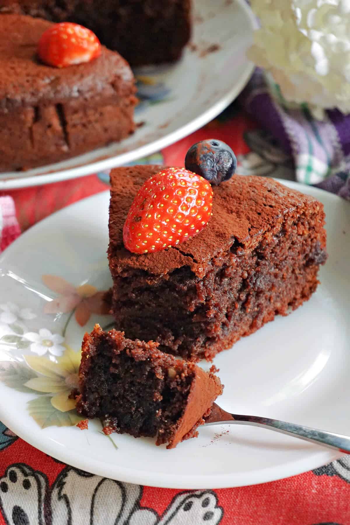 A white flowery plate with a slice of chocolate cake topped with half a strawberry and a blueberry. A fork cuts through the cake. Partially-visible plate with more cake at the top.