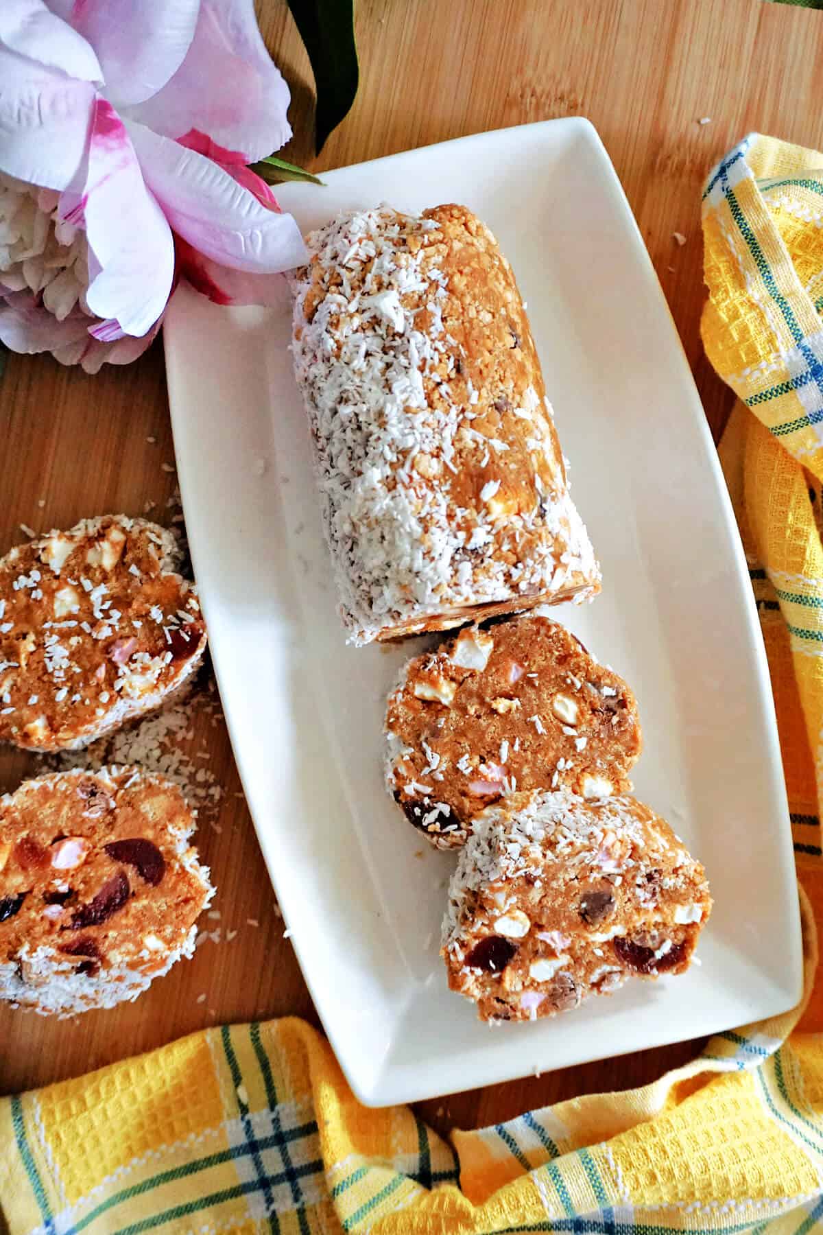 A white rectangle plate with a cake log and 2 slices of cake, 2 slices on the side, partially-visible yellow tea towel and flowers.