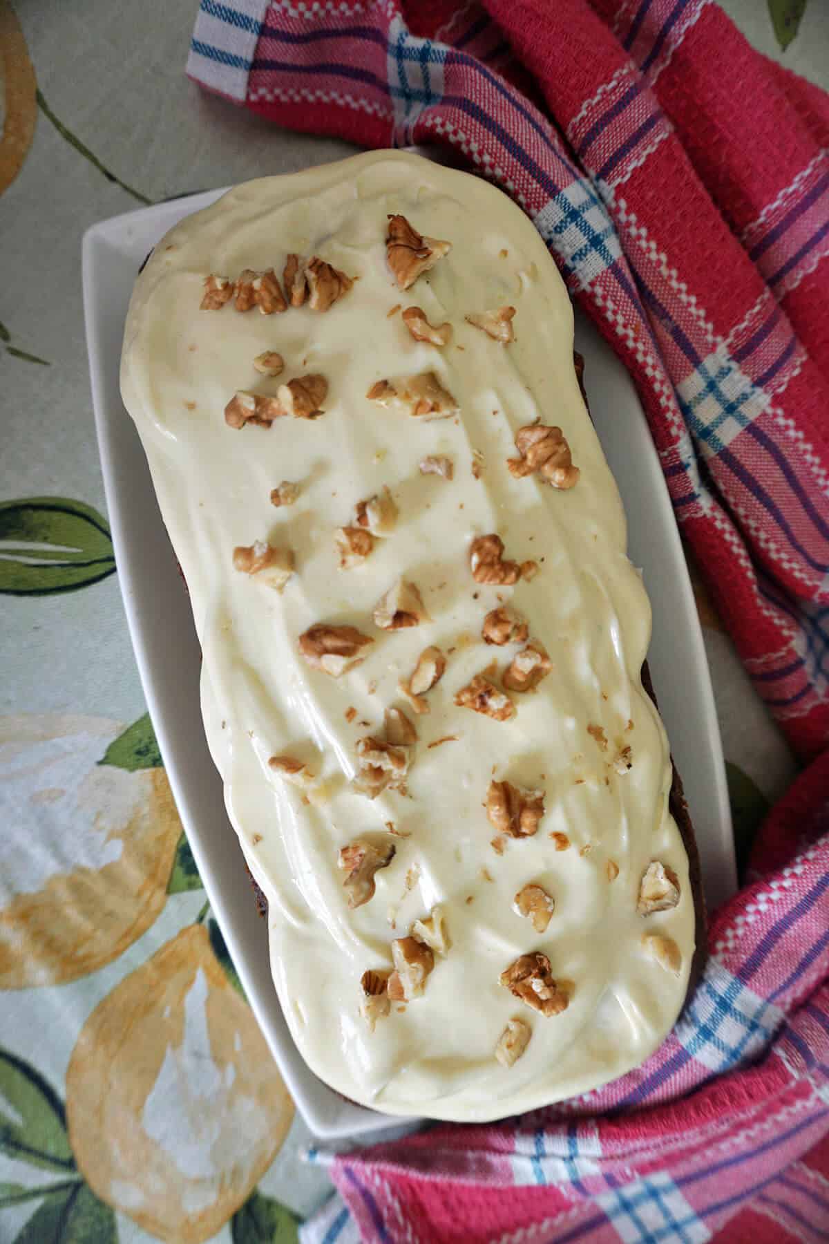 A loaf tin with a cake topped with frosting and chopped walnuts. A pink-patterned tea towel around the tin.
