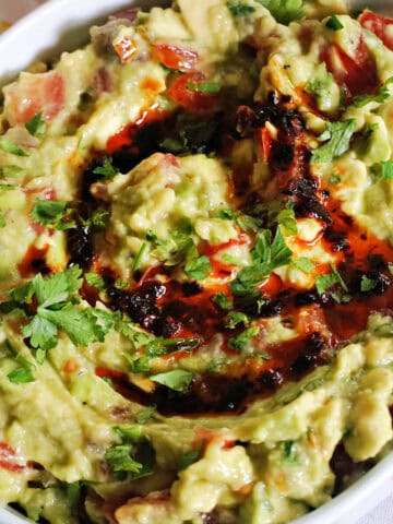A white serving bowl with chunky guacamole drizzled with chilli oil and garnished with chopped parsley.