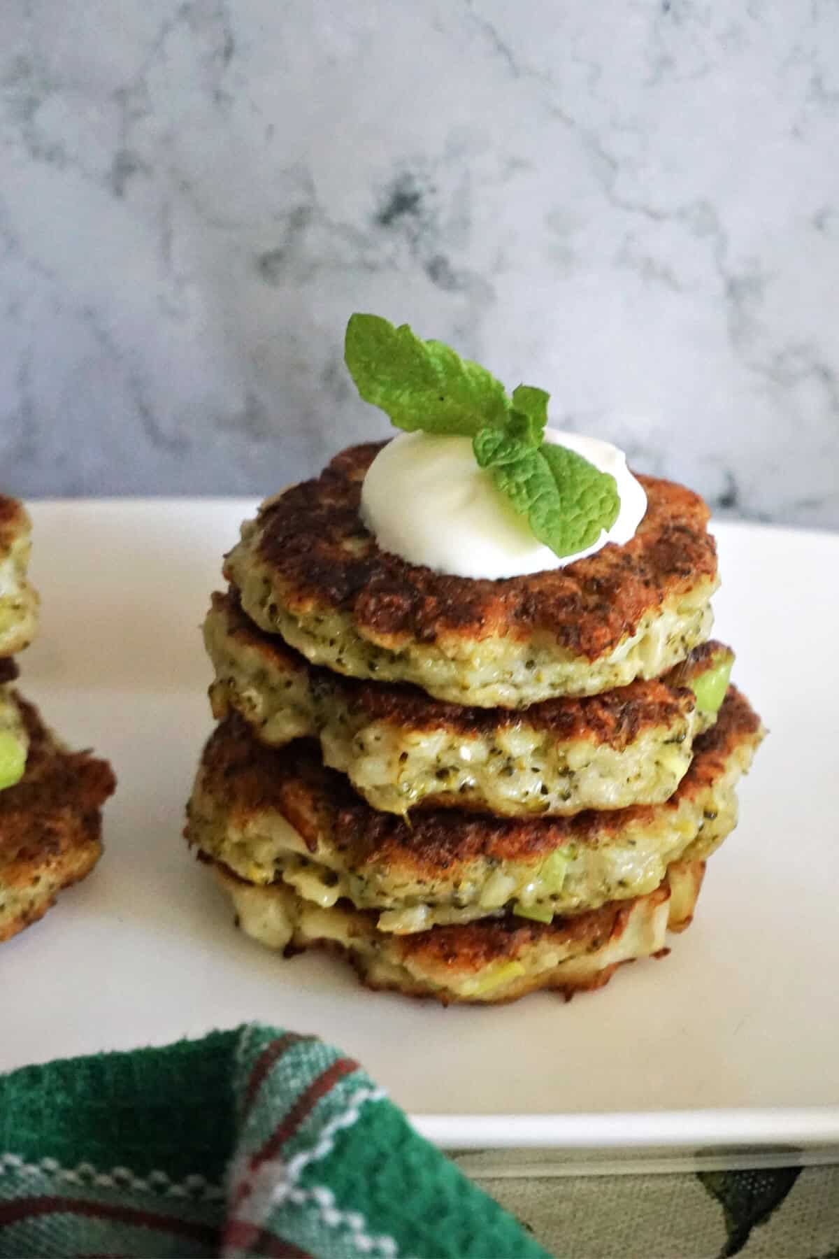 A stack of 4 broccoli patties on a white plate topped with yogurt and mint leaves.