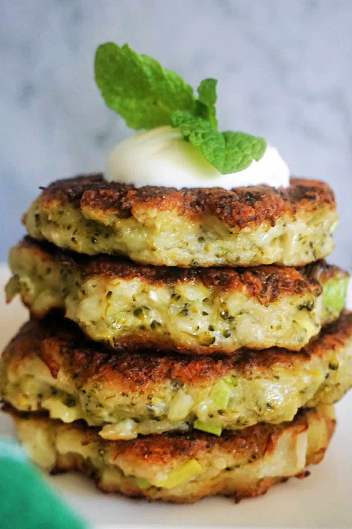 A stack of 4 broccoli fritters topped with yogurt and fresh mint leaves.