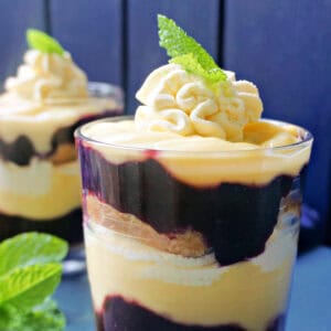 A glass with trifle topped with mint leaves, fresh mint leaves on the side and a partially-visible glass with trifle in the background.