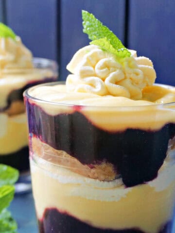 A glass with trifle topped with mint leaves, fresh mint leaves on the side and a partially-visible glass with trifle in the background.
