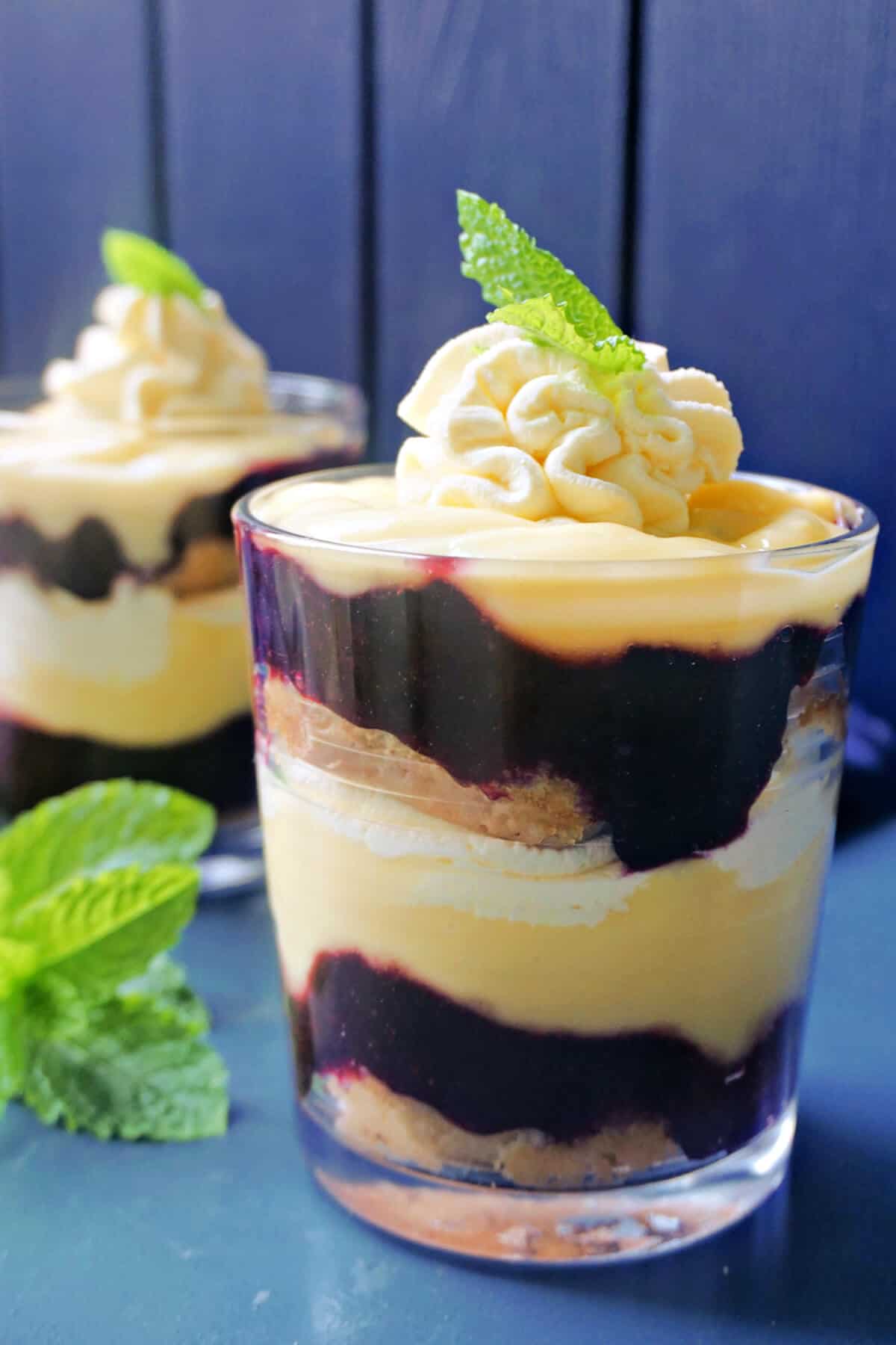 A glass with berry trifle, fresh mint leaves around it and a partially-visible glass with trifle in the background.