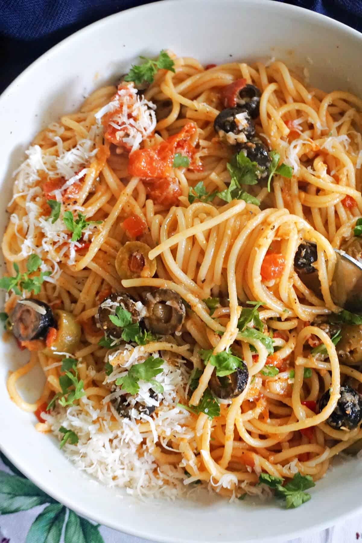 A white bowl with spaghetti puttanesca garnished with grated parmesan and chopped parsley.