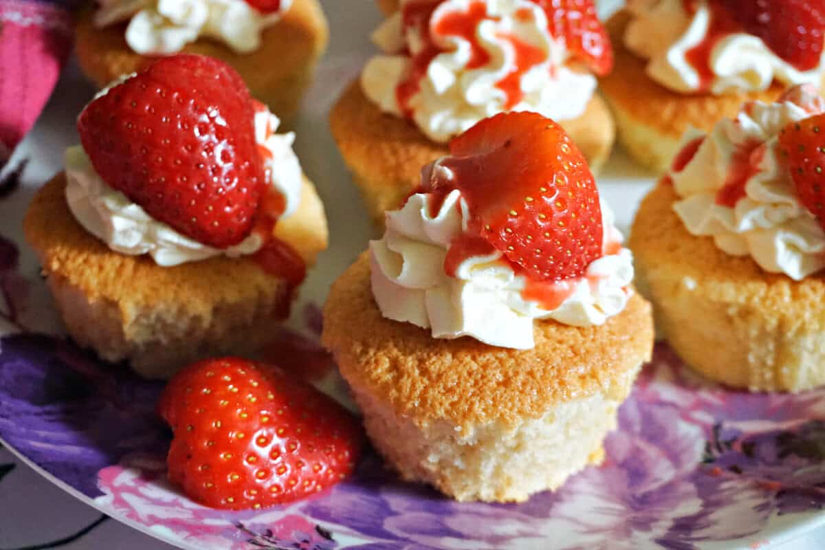 Cupcakes topped with cream, strawberry sauce and fresh strawberry halves on a white and purple plate.