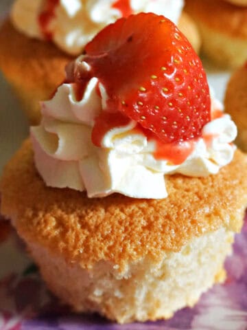 A cupcake topped with cream, strawberry sauce and half a strawberry. Other partially-visible cupcakes around it.
