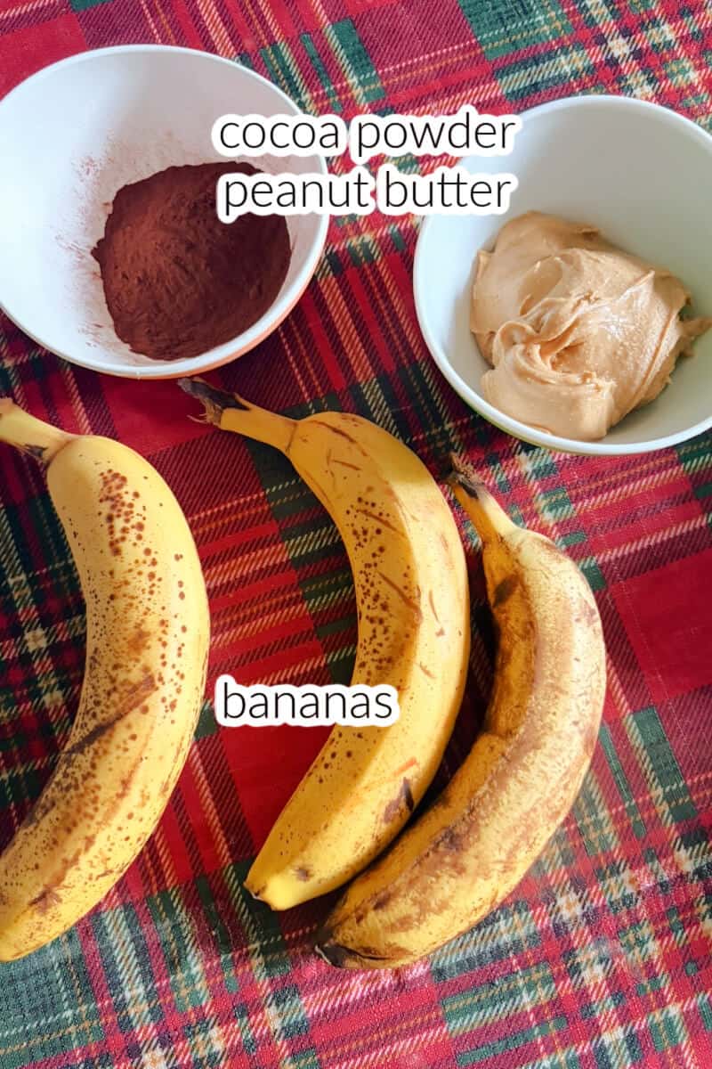 A small ramekin with peanut butter, another one with cocoa powder and 3 ripe bananas on a tablecloth.