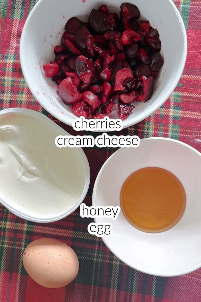 A small ramekin with honey, another one with chopped cherries, one egg and a tub of cream cheese on a tabletop.