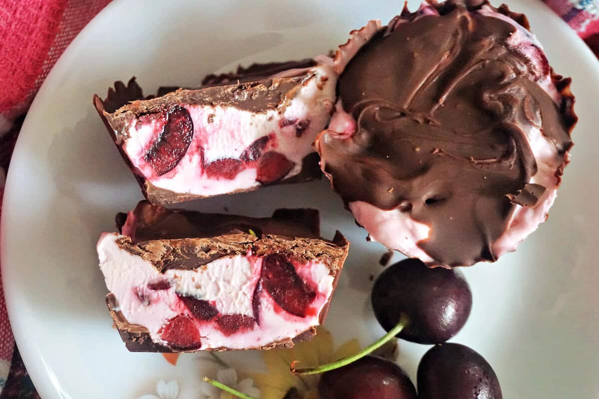A white flowery plate with 2 halves of a cherry cheesecake cup, 3 cherries and a whole chocolate-covered cup next to them.