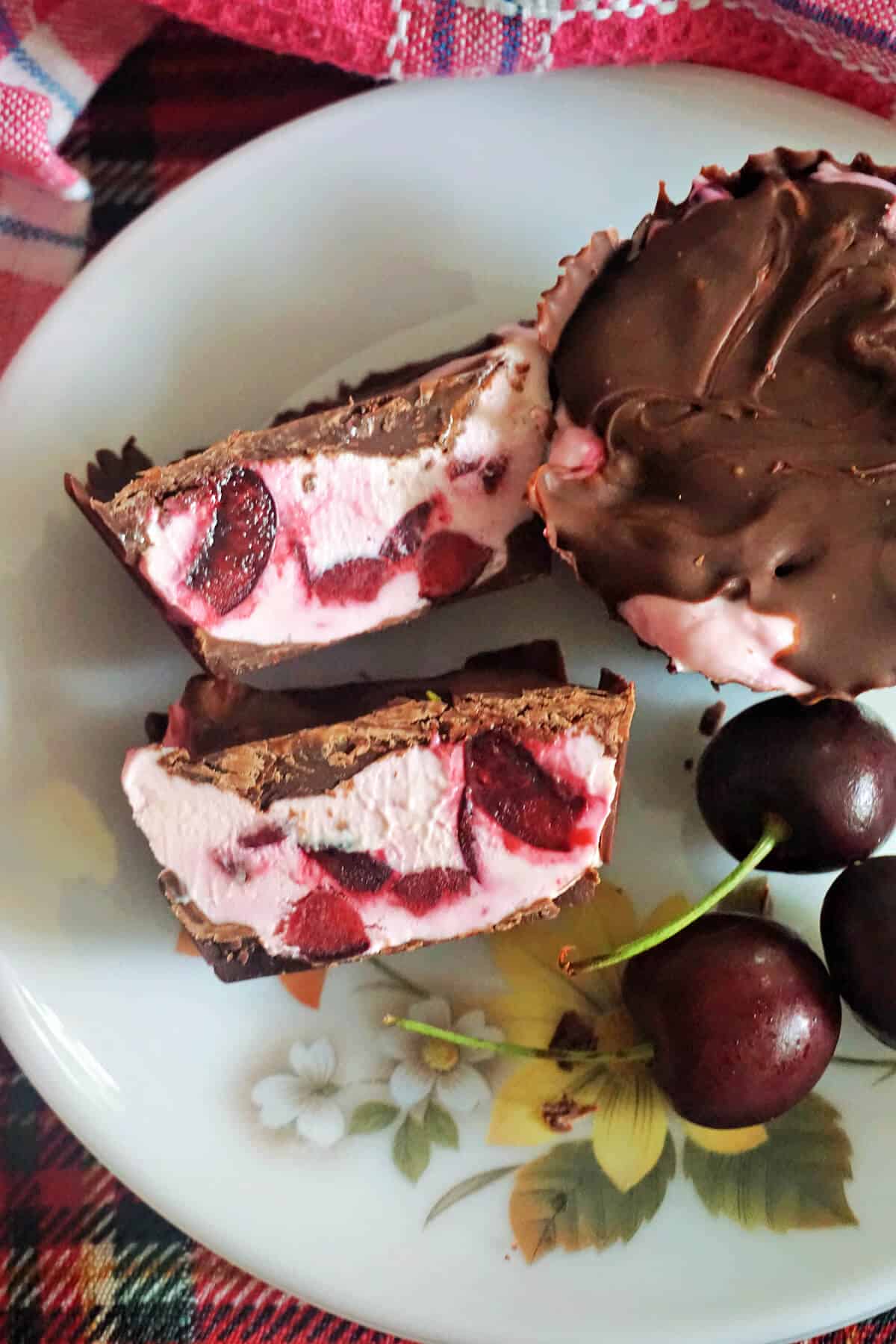 A white flowery plate with 2 halves of a cherry cheesecake cup, 3 cherries and a partially-visible whole chocolate cup.