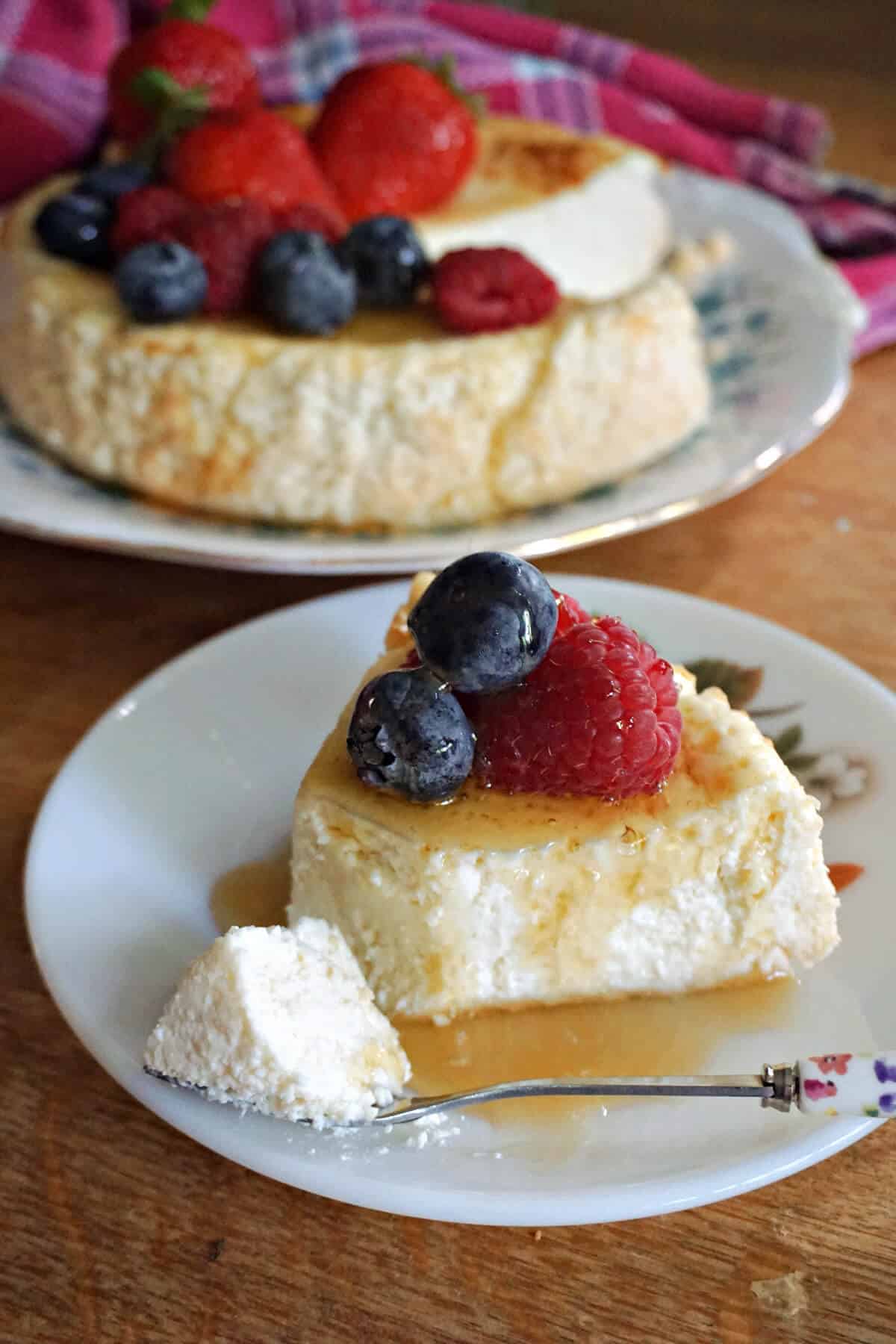A white dessert plate with a slice of crustless cheesecake topped with 2 blueberries and a raspberry and drizzled with honey. A fork cutting through it. A partially-visible plate with the rest of the cheesecake in the background.