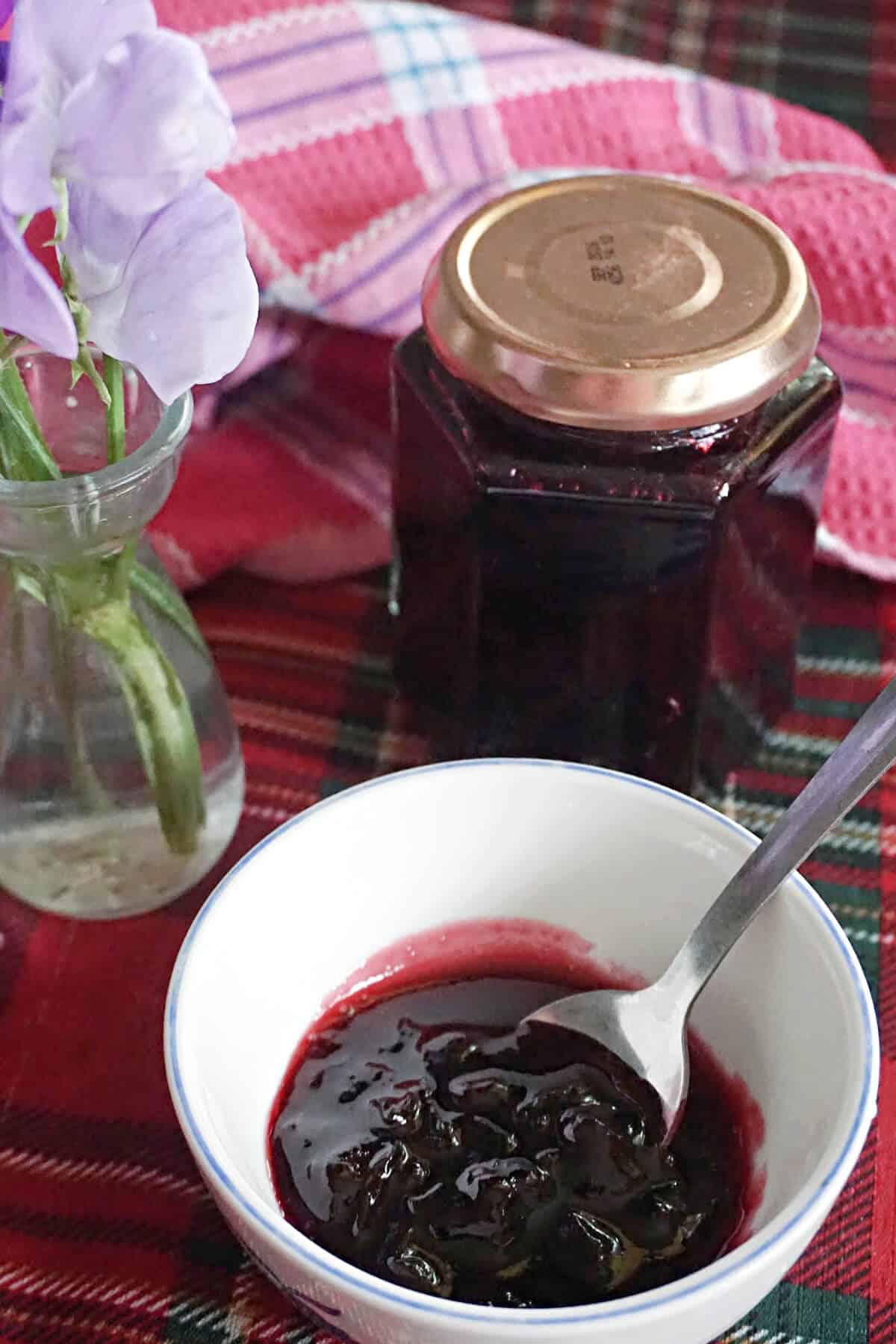 A spoon in a small bowl with jam, a jar of jam at the top with a partially-visible small vase of flowers next to it.