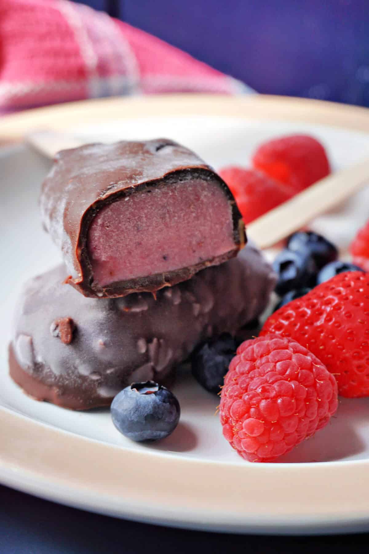 A white plate with half a popsicle on top of a whole popsicle and fresh berries around.