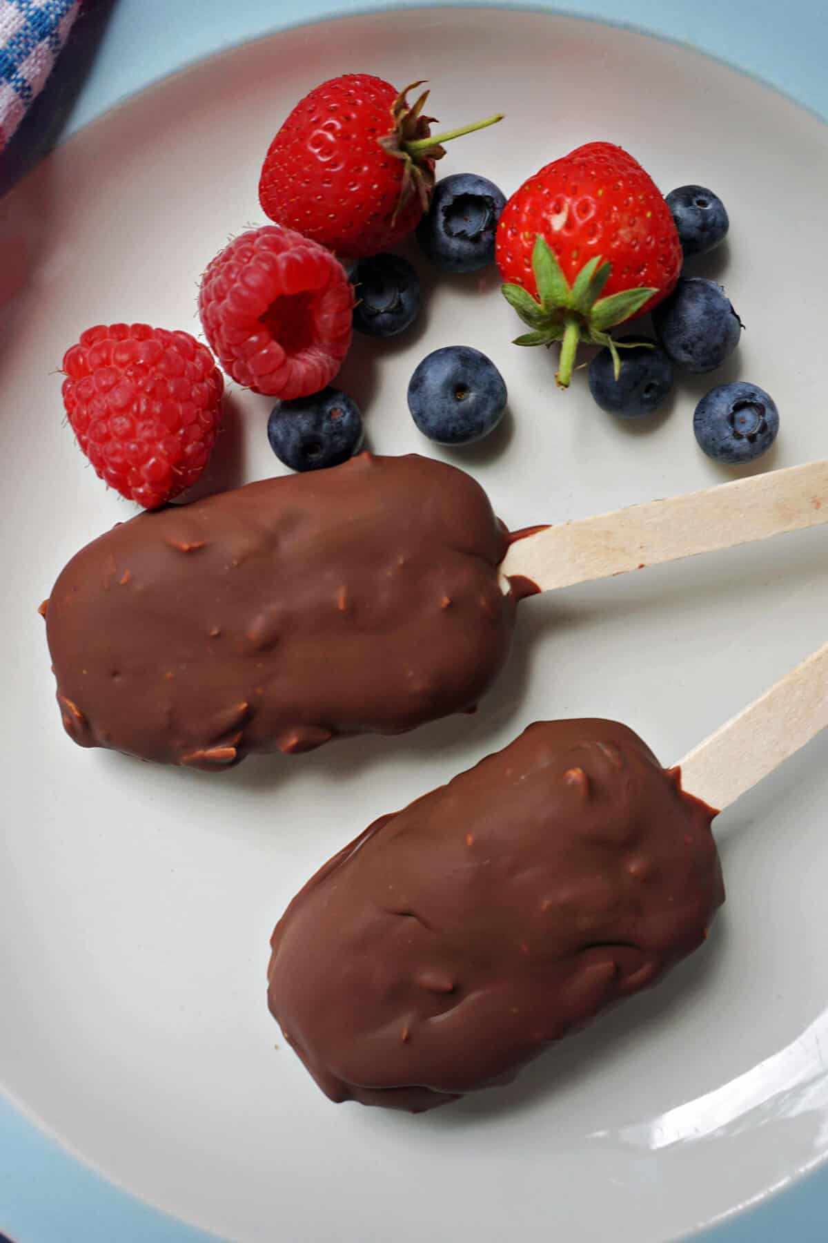 A white plate with 2 chocolate-covered ice creams and fresh berries around them.