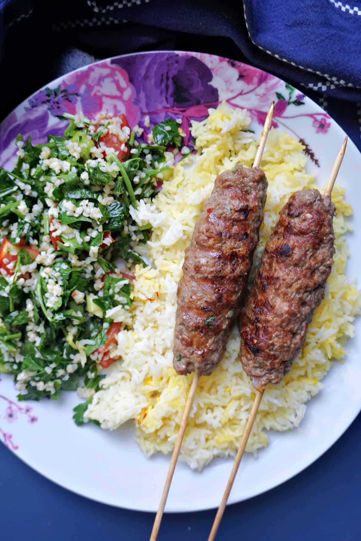 A plate with 2 kofta kebab skewers over a bed of gold rice and salad on the side.