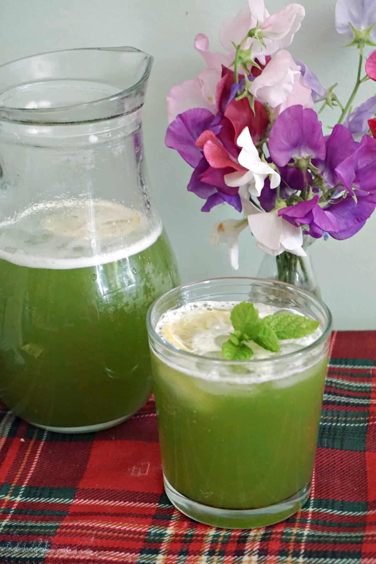A glass of green lemonade and a jug of lemonade next to it. A partially visible small base of flowers in the background.