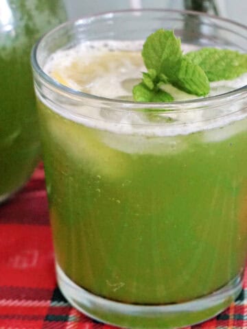 A glass of green lemonade garnished with fresh mint leaves. A partially-visible jug of lemonade next to it.